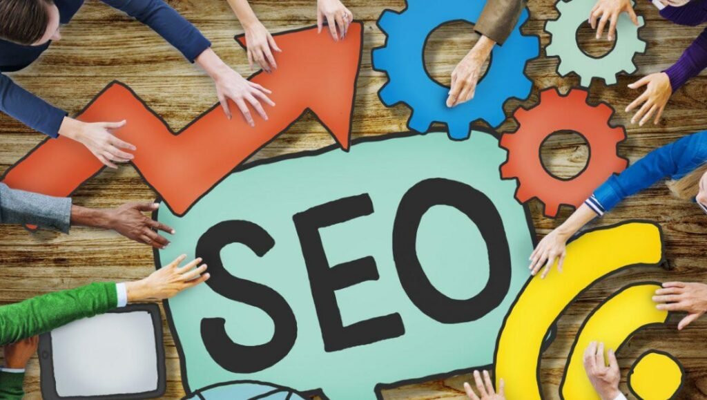 Overhead view of several hands arranging large colorful letters and shapes to spell "SEO" on a wooden background, surrounded by multicolored cogwheels, symbolizing teamwork and search engine optimization.