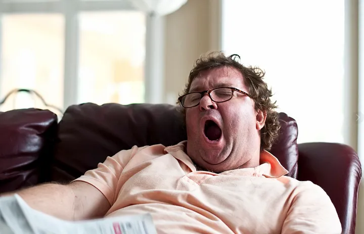 Man in pink shirt yawning on leather sofa, holding newspaper in well-lit living room.