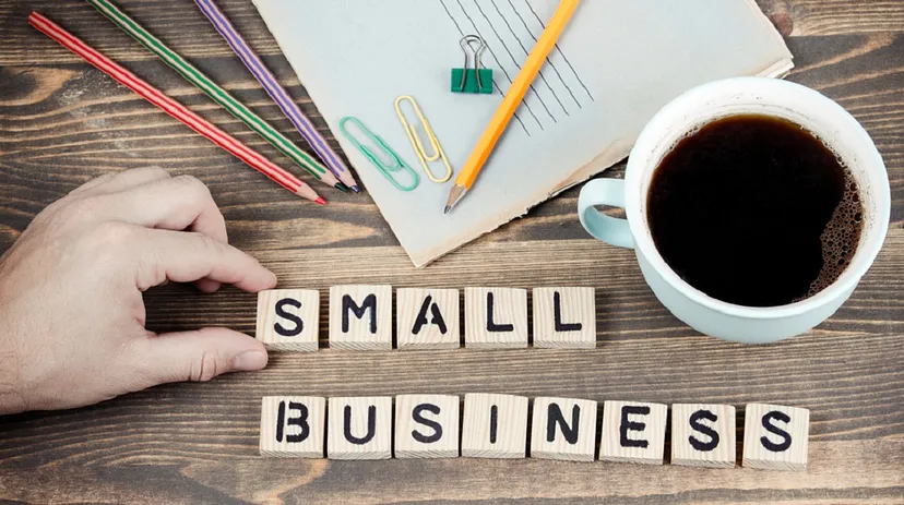 A hand resting near wooden blocks spelling "SMALL BUSINESS" on a desk with a cup of coffee, paper, colourful pencils, and paper clips.