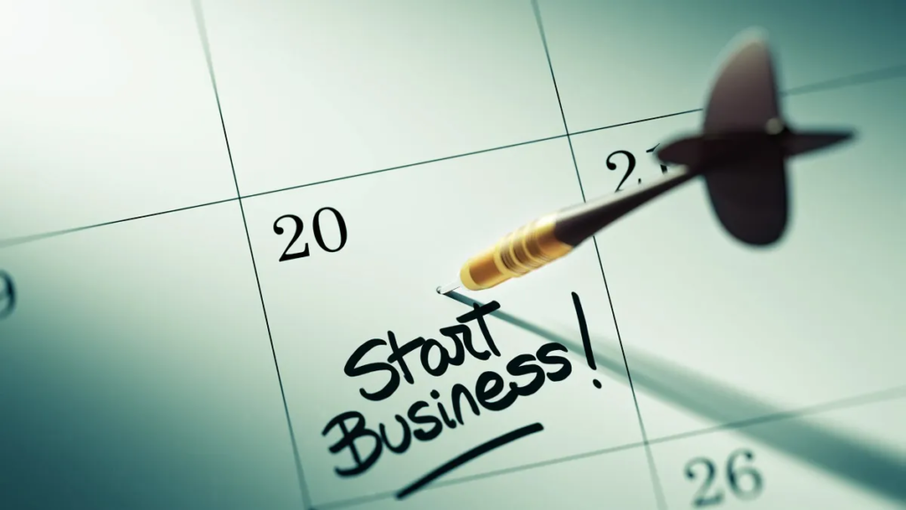 A dart pinned to a calendar on the date 23 with the words "Start Business!" handwritten on it.