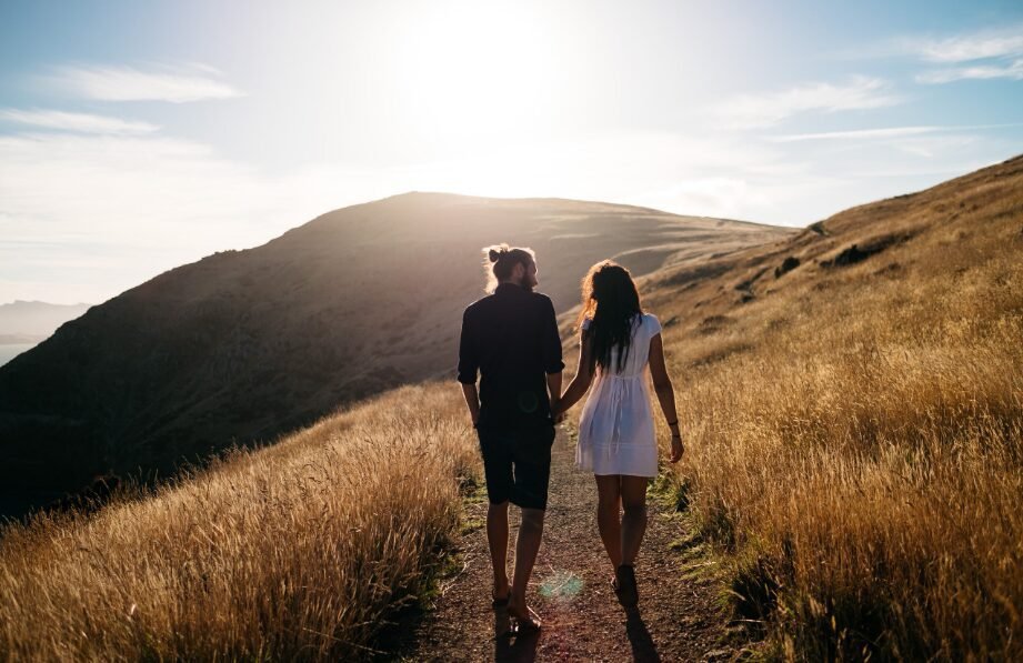 A couple holding hands and walking through a sunlit grassy field with rolling hills in the background.