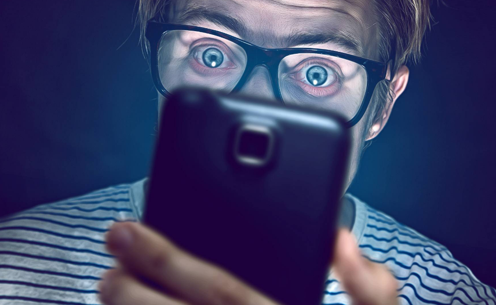 A close-up of a surprised person with glasses holding a smartphone camera towards the viewer.