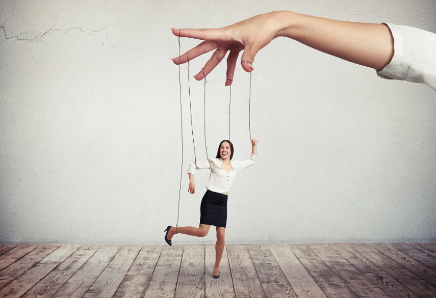 A woman in business attire appears to be a marionette being manipulated by a large hand using strings against a plain wall and wooden floor background.