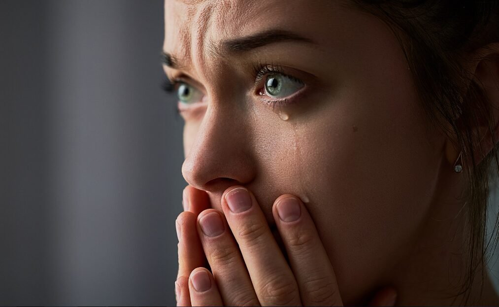Distressed woman with tear-filled eyes and hands covering mouth in emotional turmoil.