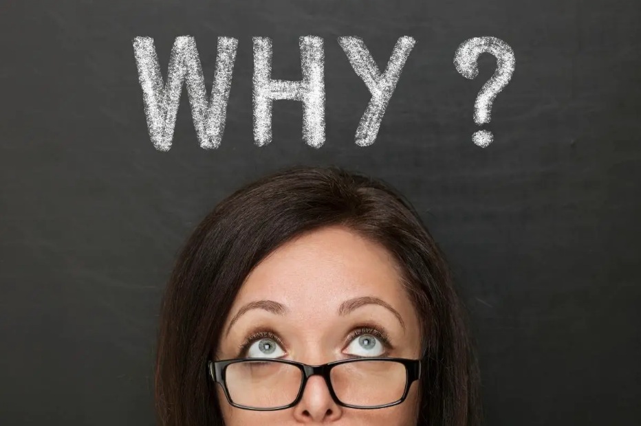 A woman with glasses looking upwards with a thoughtful expression, standing in front of a blackboard with the word "WHY?" written in white chalk above her head.