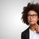 Professional woman in afro hairstyle and black blazer gazes thoughtfully against gray backdrop.