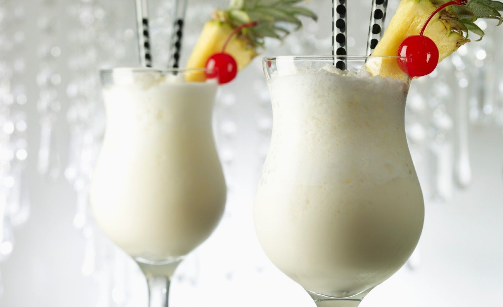 Two pina colada cocktails in hurricane glasses, garnished with pineapple slices and maraschino cherries, with black straws, against a blurred background of champagne flutes.