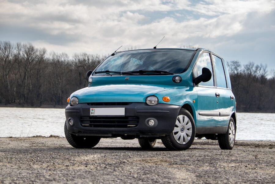 A turquoise Fiat Multipla parked on a gravel surface with leafless trees and a calm river in the background under a cloudy sky.