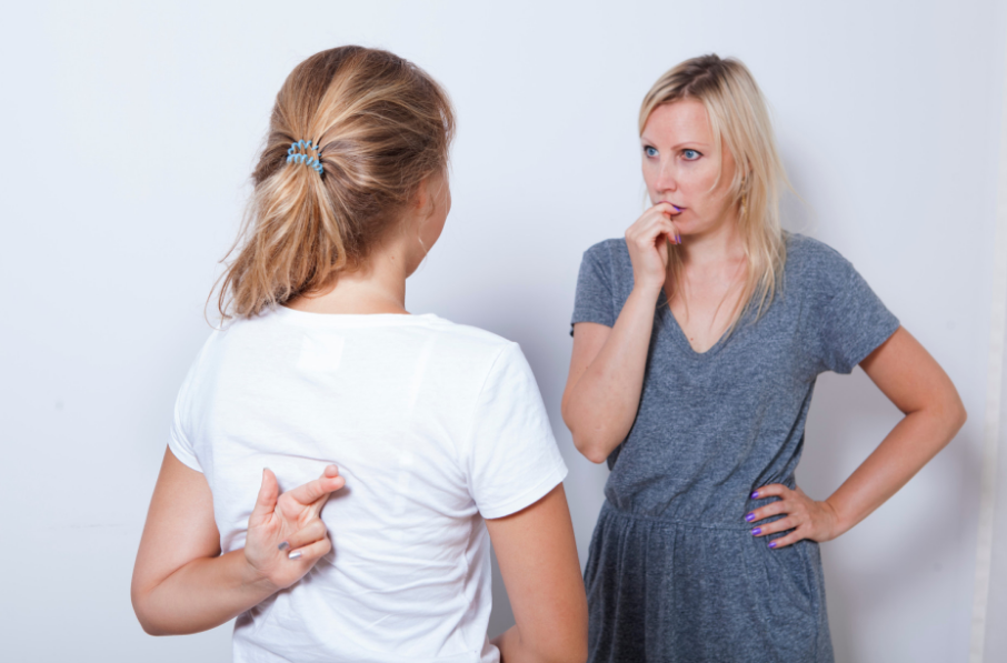 Two women having a conversation; one is gesturing behind her back with her fingers crossed, indicating deception or a lie, while the other looks on with a suspicious expression.