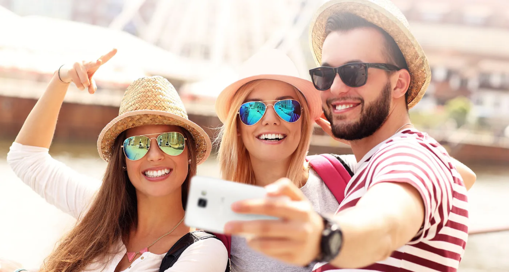 Three young adults wearing summer hats and sunglasses taking a selfie with a smartphone, smiling, and posing in front of a bright waterfront scene.