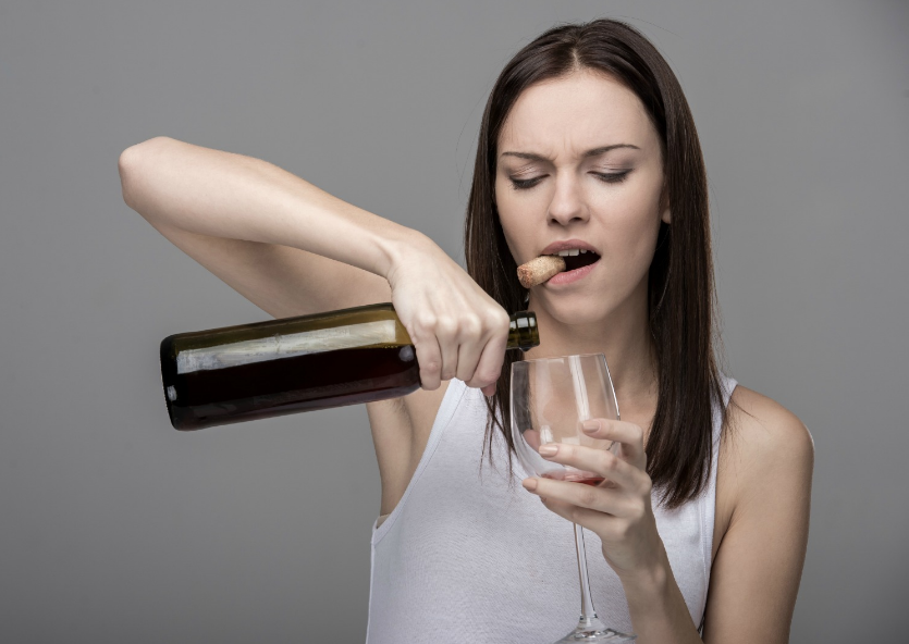 A woman in a white tank top struggling to open a wine bottle with her teeth while holding a wine glass in her other hand against a grey background.