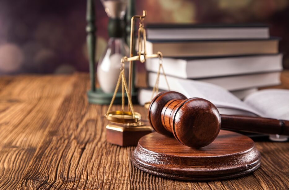 A wooden judge's gavel and sound block rest on a table with a brass scale of justice and a stack of books in the background, suggesting a legal theme.