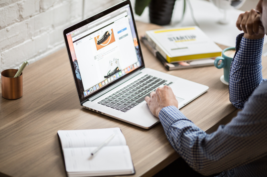 A person working on a laptop with a website displayed on the screen, with a notepad and pen, and a cup of coffee on the desk. They are wearing a blue checked shirt. There is a plant and a stack of books in the background.