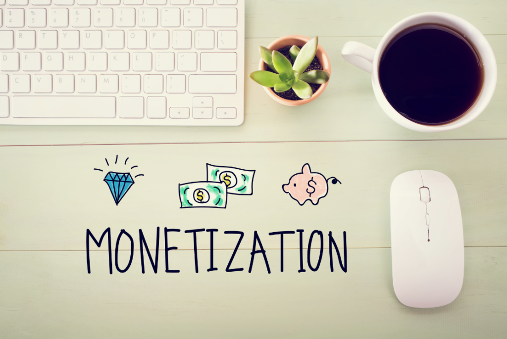 A workspace with a white keyboard and mouse, a potted plant, a cup of coffee, and hand-drawn illustrations of a diamond, money, a piggy bank, and the word "MONETIZATION" on a wooden desk.