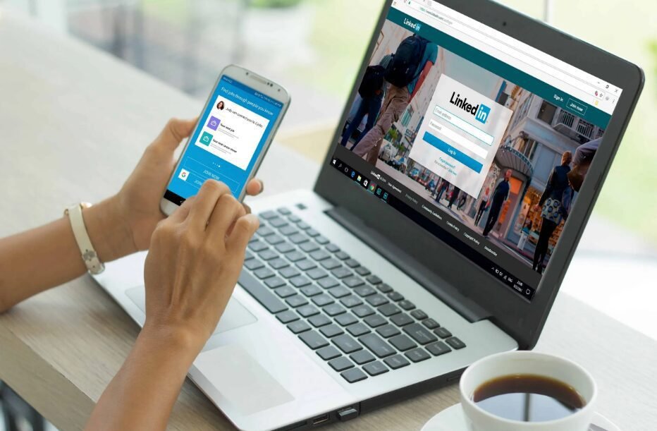 Person using a smartphone with a social media app open in front of a laptop displaying a professional networking website's login page, with a cup of coffee on the side.