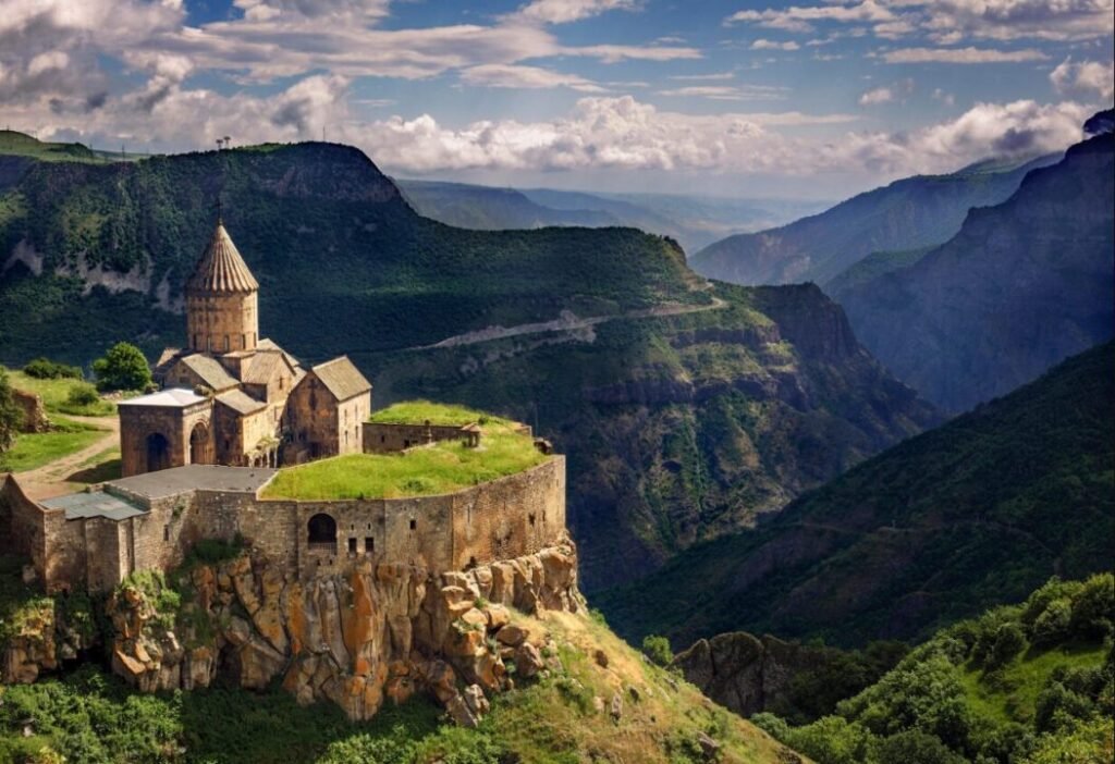 Ancient fortress with a stone church on a cliff overlooking a mountainous landscape with lush greenery and distant clouds.