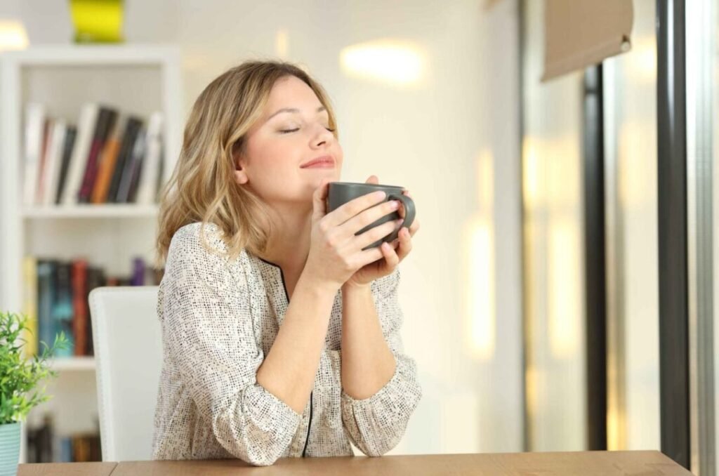 Woman enjoying the aroma of a hot drink from a mug, sitting at a table in a brightly lit room with bookshelves in the background.