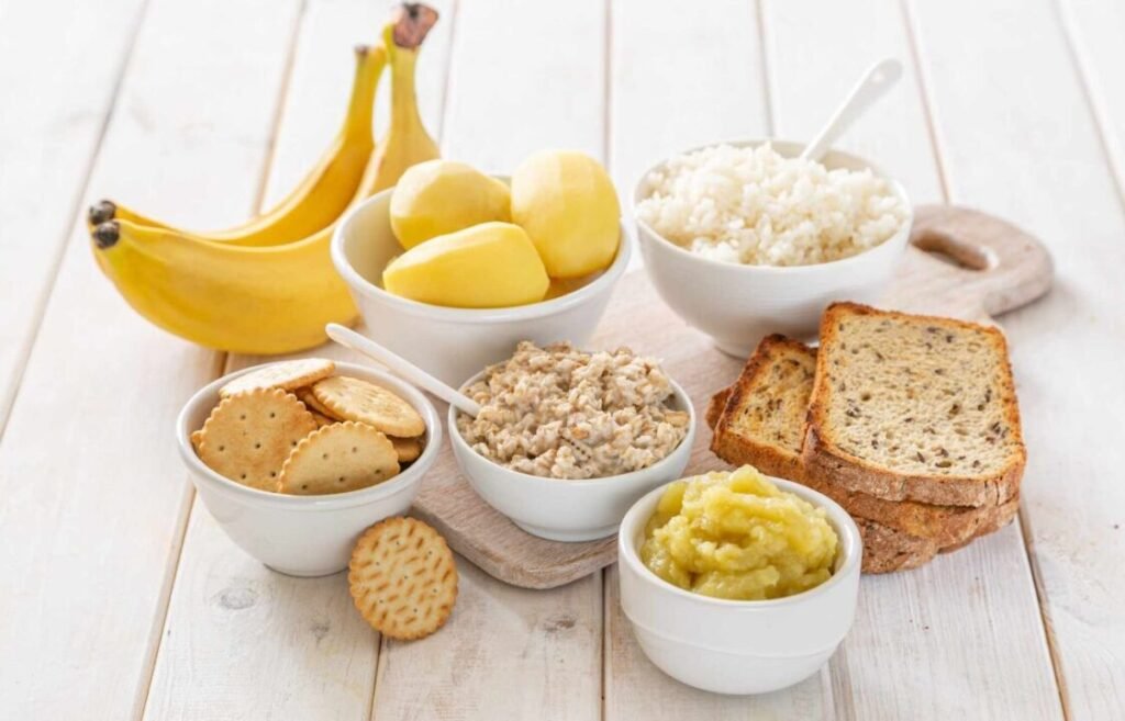 Assorted carbohydrate-rich foods on a wooden table, including bananas, peeled potatoes, white rice, oatmeal, crackers, whole grain bread, and applesauce.