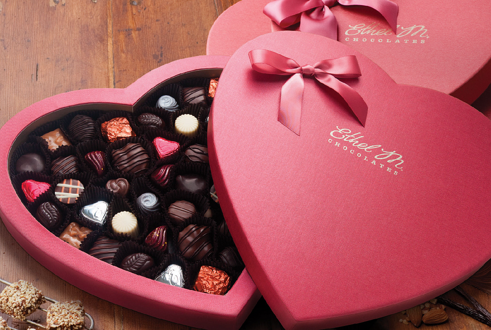 Two heart-shaped boxes of Ethel M Chocolates, one open to show assorted chocolates, on a wooden table.