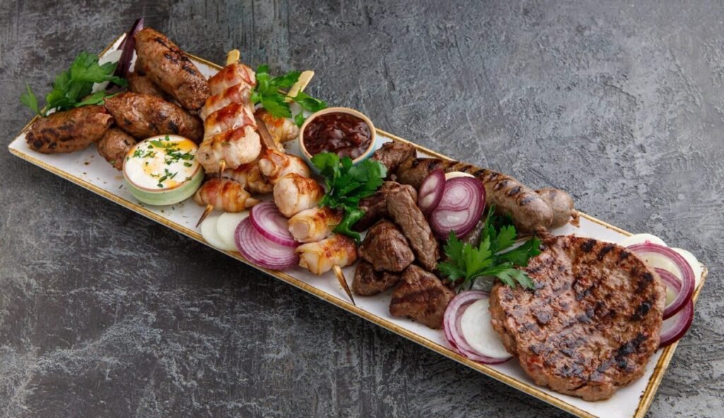 A platter of assorted grilled meats including skewers of shish kebab, sausages, and burgers, accompanied by dips and garnished with parsley and red onion slices on a long rectangular plate, set against a dark textured background.