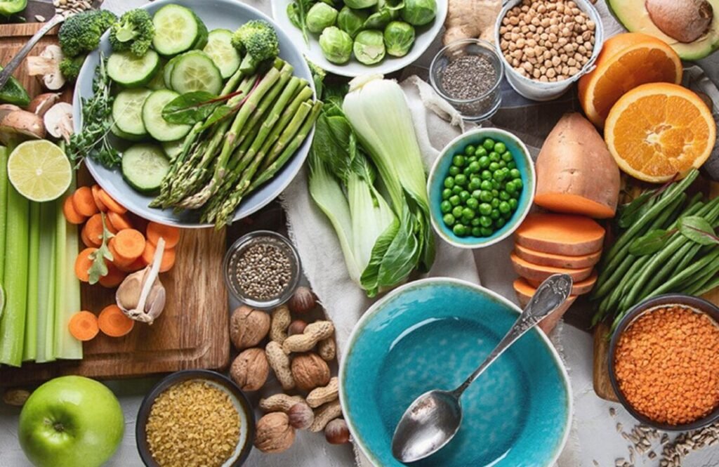 Assorted fresh vegetables, legumes, nuts, and grains spread out on a table, including cucumbers, carrots, asparagus, peas, sweet potato, and citrus fruits, representing a healthy, plant-based diet.