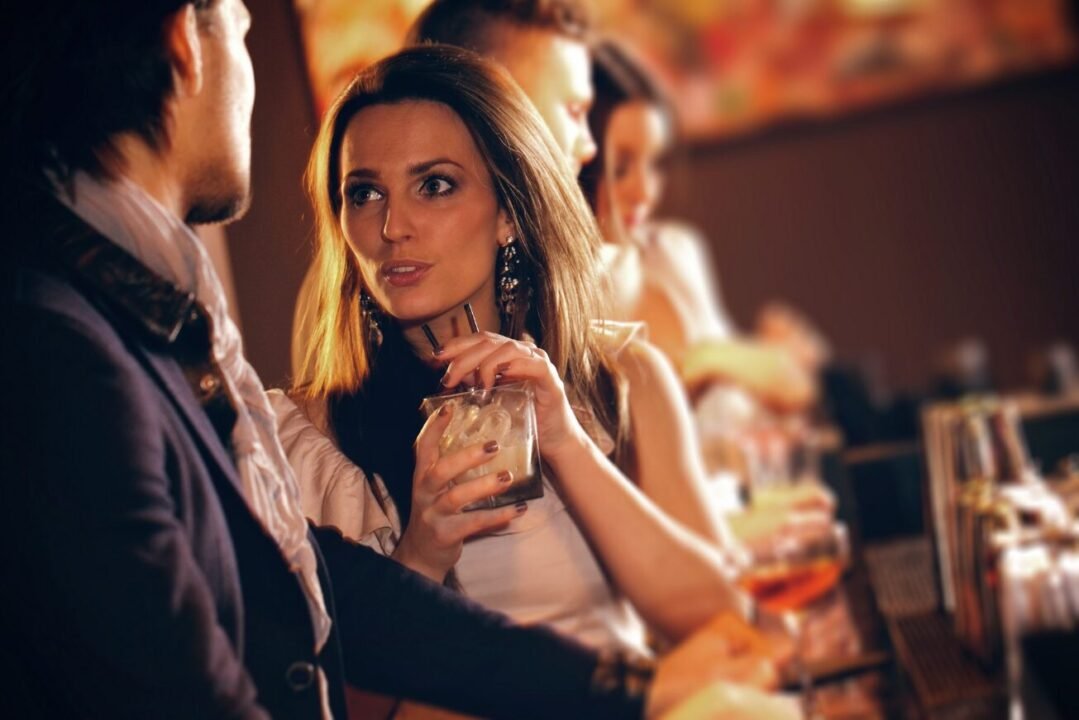 Couple conversing in warm bar setting, others blurred in background.