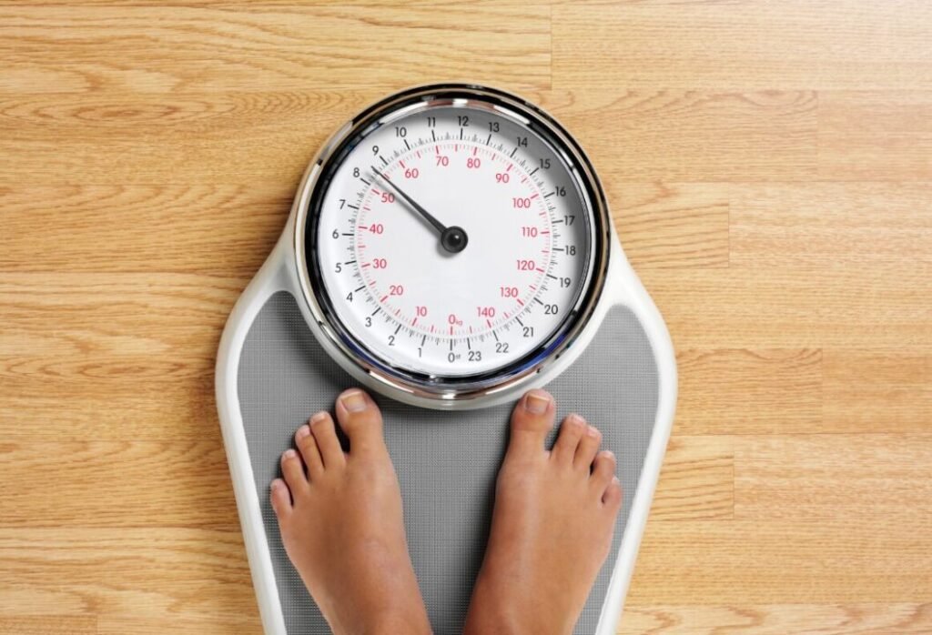 A person's bare feet standing on a mechanical bathroom scale with a dial indicator on a wooden floor.