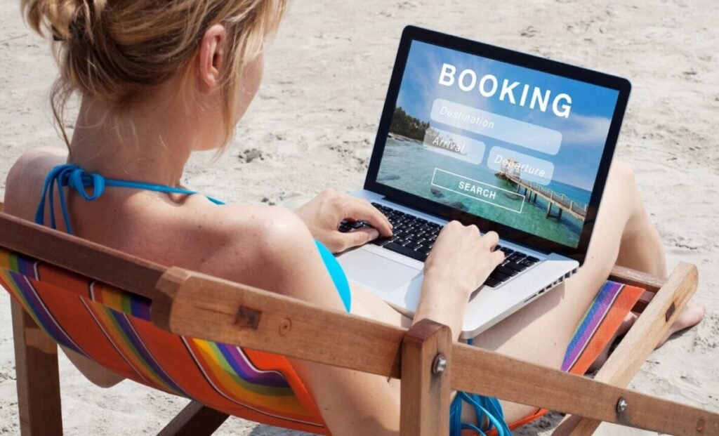 Woman in a colorful beach chair using a laptop with a travel booking website displayed on the screen, at the beach.