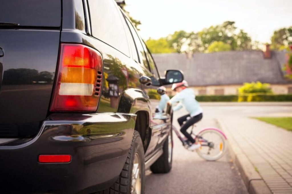 A young girl wearing a helmet riding a bike closely passes by a parked black SUV on a sunny suburban street.