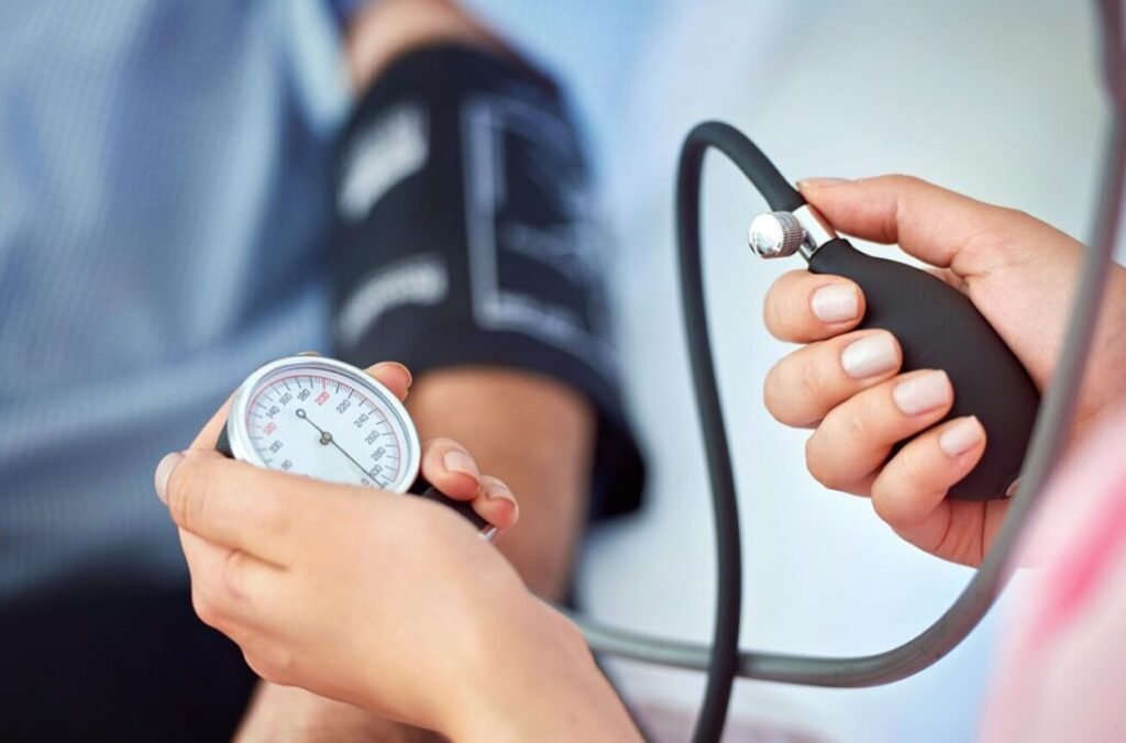 A healthcare provider is taking a patient's blood pressure, showing a sphygmomanometer gauge in focus with the cuff wrapped around the patient's arm in the background.