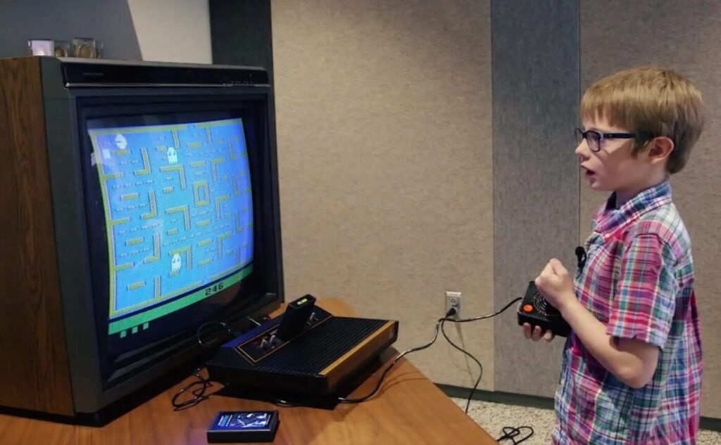 A young boy in glasses and a plaid shirt playing a classic video game on an old-fashioned home console connected to a large CRT television displaying a pixelated game screen.