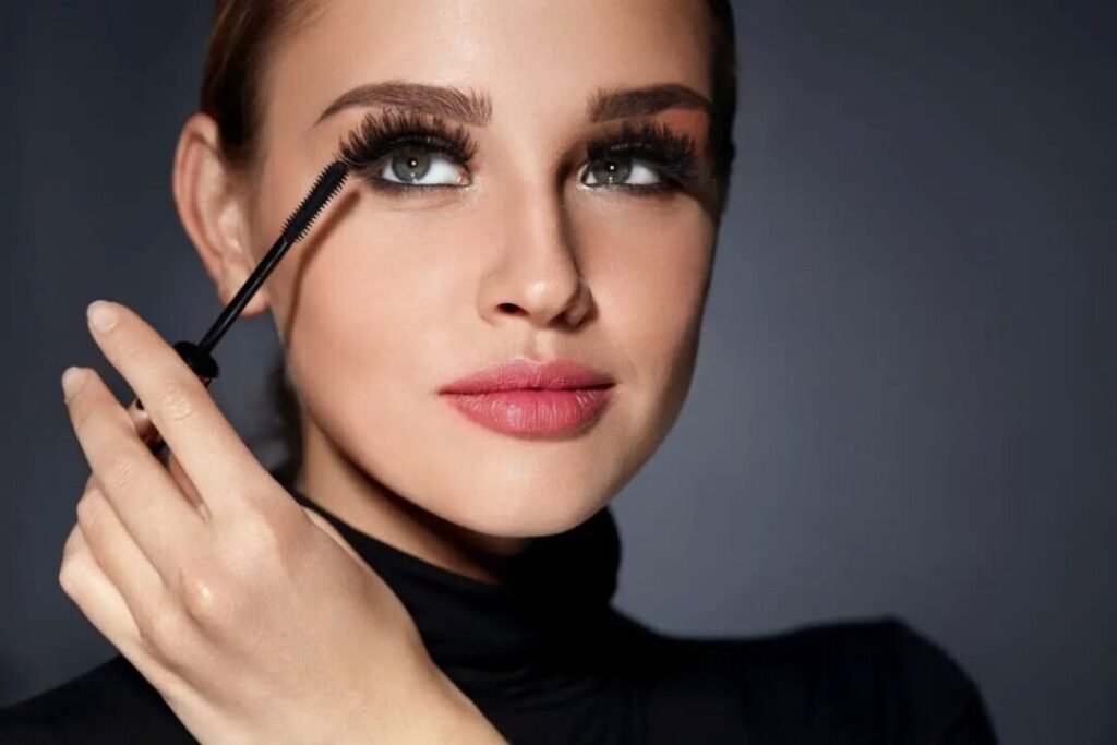 Woman applying mascara to her eyelashes, wearing a black turtleneck, against a grey background.