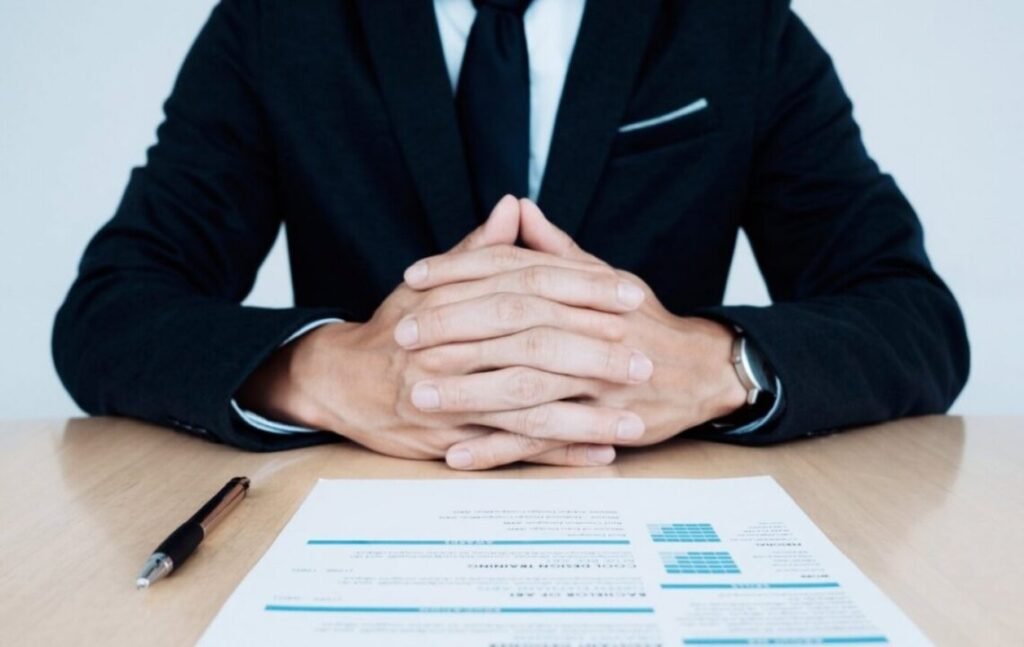 Businessman in a suit sitting at a desk with his hands clasped, documents and a pen in front of him.