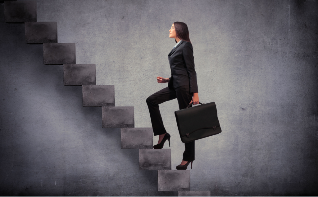 A businesswoman in a black suit and heels carrying a briefcase while ascending abstract stone steps against a textured grey background.