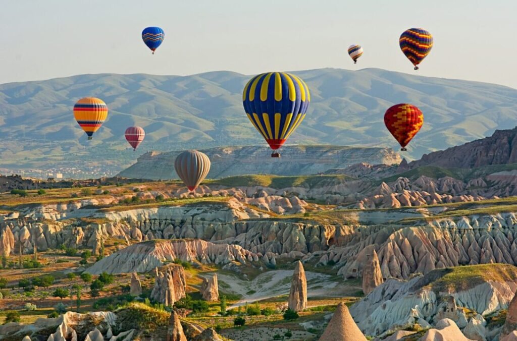 Multiple hot air balloons floating over a rocky landscape with distinctive geological formations and distant mountains under a clear sky.