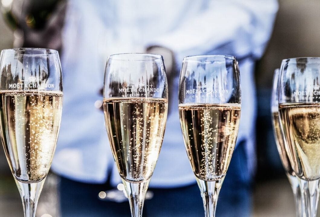 Four champagne glasses filled with sparkling champagne, branded with "Moët & Chandon", with a blurred waiter in the background.