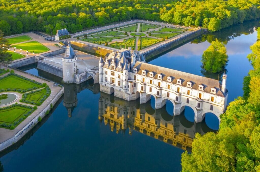 Aerial view of the Château de Chenonceau, a French Renaissance castle spanning the River Cher, with formal gardens and reflective water on a sunny day.