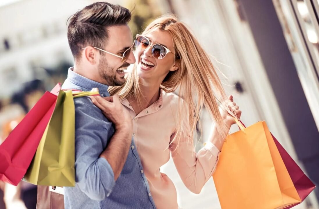 A cheerful couple wearing sunglasses and carrying colorful shopping bags, hugging and smiling joyfully at each other on a sunny day outdoors.