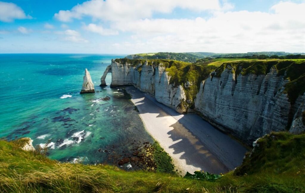 A scenic view of Etretat's white chalk cliffs and arches along the blue waters of the English Channel, with a natural arch detached from the main cliffs, a pebble beach below, and lush greenery atop the cliffs under a partly cloudy sky.