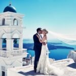 Bride and groom embrace by white bell tower overlooking blue Aegean Sea.