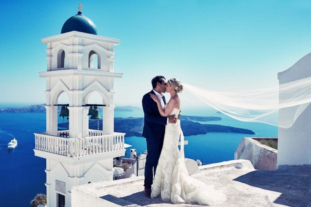 Bride and groom embrace by white bell tower overlooking blue Aegean Sea.