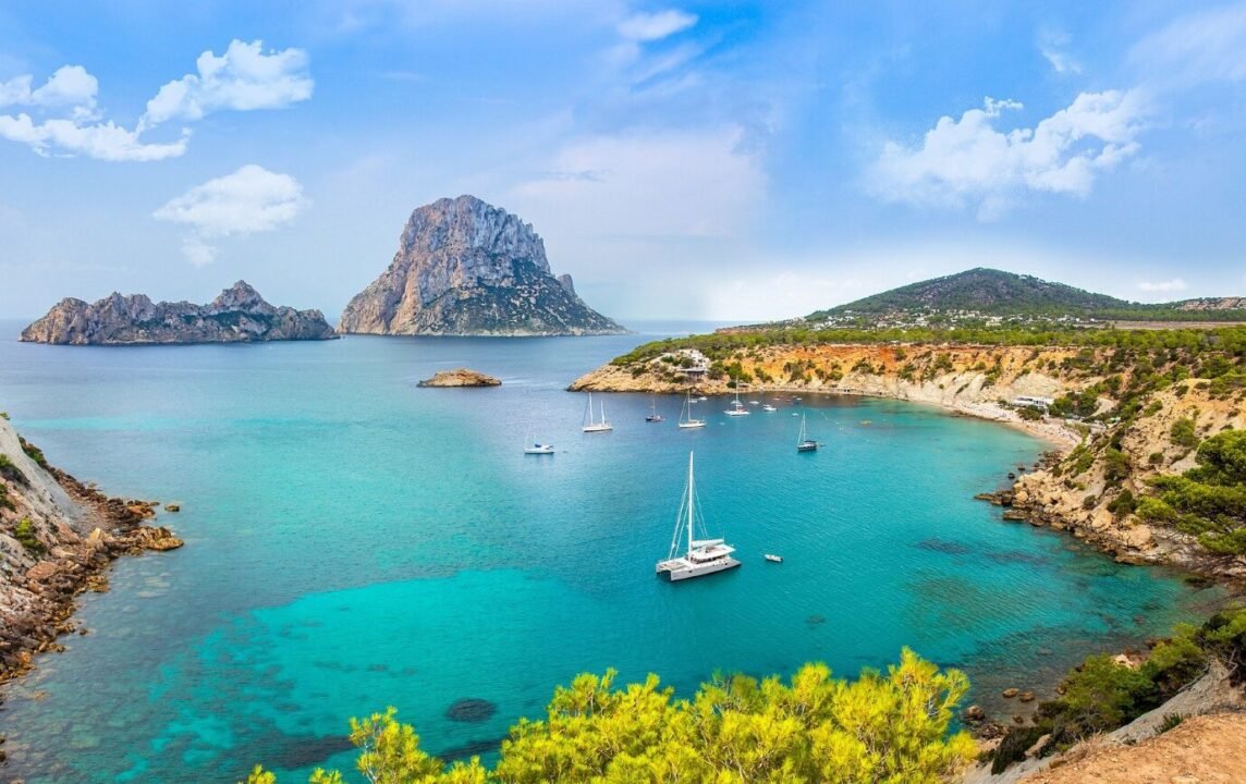 A panoramic view of a coastal landscape featuring a clear turquoise sea with several sailboats, a prominent rocky mountain in the background, and lush greenery along the coastline under a partly cloudy sky.