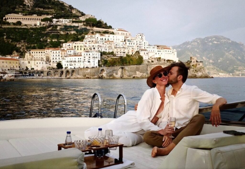 A couple enjoying each other's company on a luxury boat with a scenic view of a hillside town by the sea in the background. They are sitting close, sharing a kiss on the cheek, dressed in casual vacation attire, with a table set with drinks in the foreground.