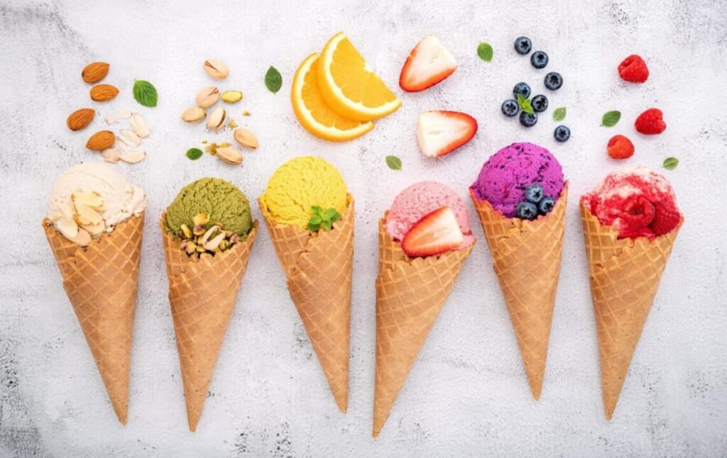 Assortment of colorful ice cream cones lined up, with flavors like pistachio, mango, and berry, garnished with nuts and fresh fruits like strawberries and oranges, on a textured white background.