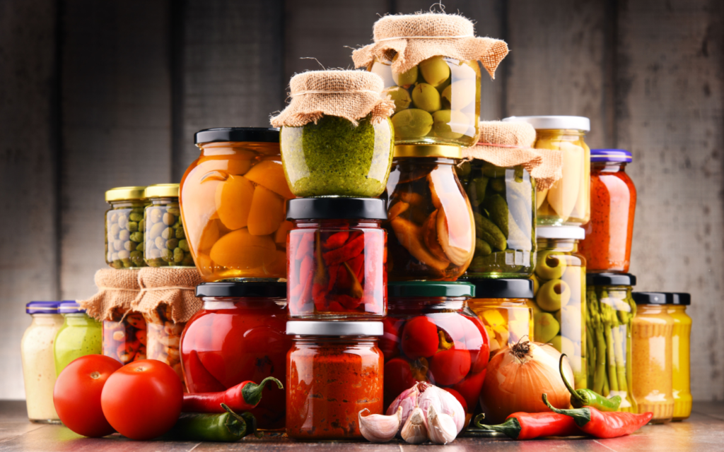 A variety of colorful pickled vegetables and sauces in glass jars, covered with burlap, presented with fresh tomatoes, chili peppers, garlic, and an onion in front of them.