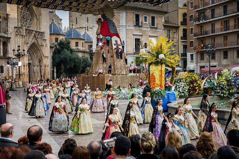 A lively festival scene with a procession of people in traditional, colorful Spanish dresses, walking in front of a large, ornate floral display with a statue. Spectators gather to watch against a backdrop of historical architecture.