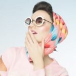 Stylish woman in retro sunglasses and colorful headscarf poses playfully against light background.
