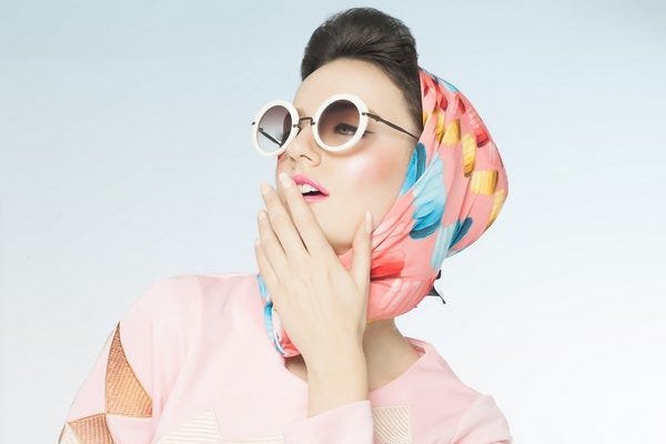 Stylish woman in retro sunglasses and colorful headscarf poses playfully against light background.