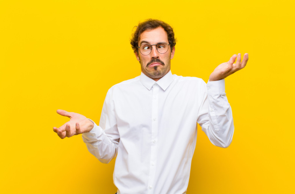 A man in a white shirt and glasses standing against a yellow background with a confused expression, shrugging his shoulders.