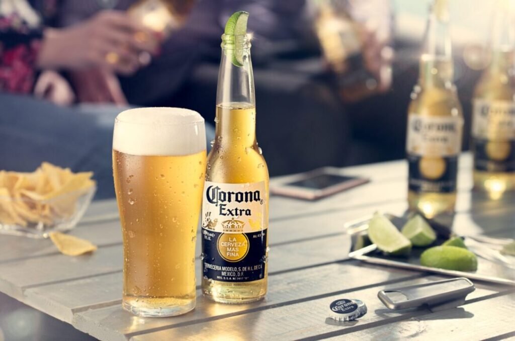 A bottle of Corona Extra beer with a lime wedge on its neck, next to a full pint glass of beer on a sunlit wooden table, with more bottles and slices of lime in the background, surrounded by people enjoying a social gathering.